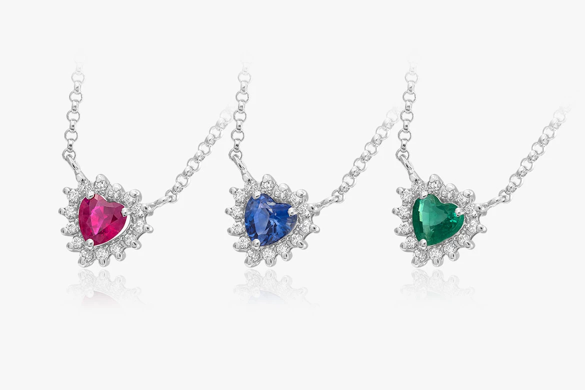 tri náhrdelníky v tvare srdca z bieleho zlata, v ktorých sú osadené diamanty, smaragd, rubín, zafír